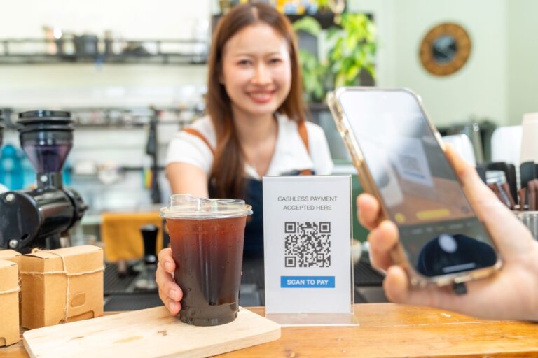 コーヒーショップのレジカウンターで、客がスマホを使ってQRコードをスキャンし、飲み物を購入している様子