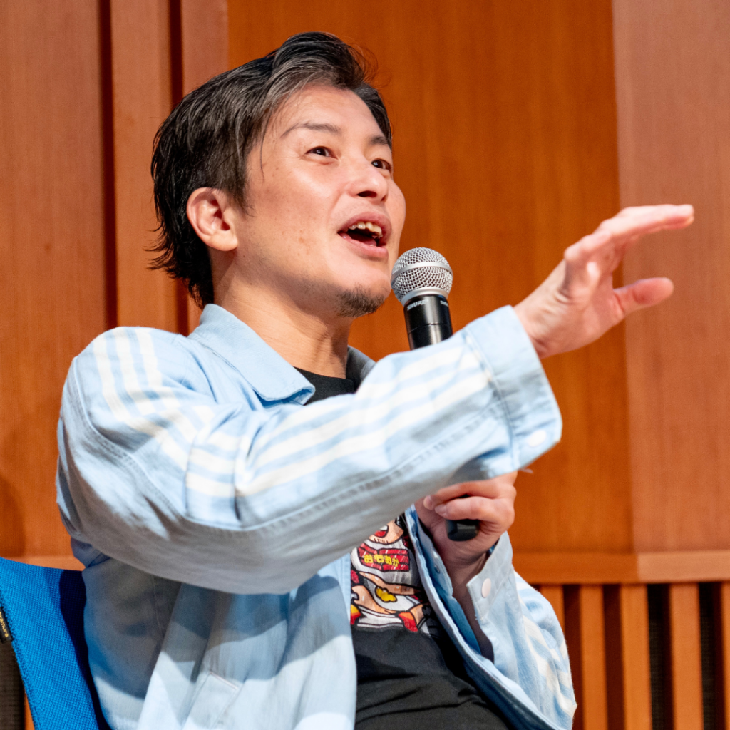 登壇者の男性が、水色の縞柄のシャツジャケット姿でマイクを持ち、手を伸ばして話す
