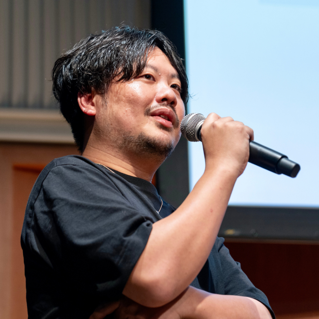 登壇者の男性が、黒いTシャツ姿でマイクを持ち、上を向いて話す