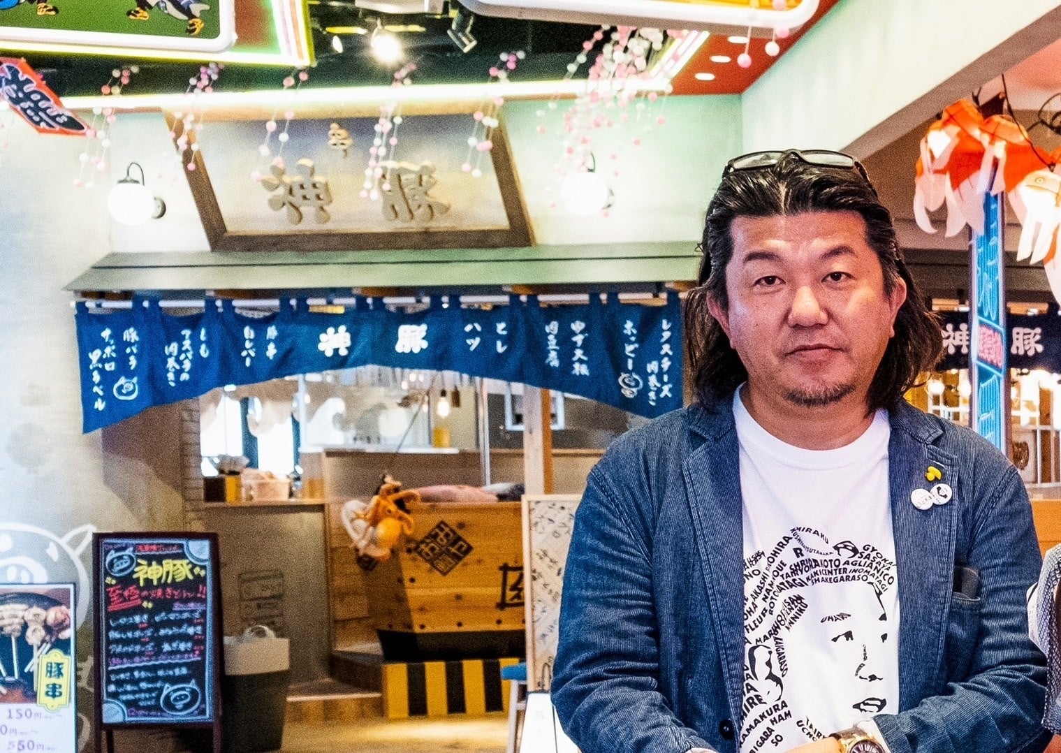 「神豚」の看板と青いのれんが並ぶ店先で、デニムのジャケットに白いTシャツ姿の男性が正面を向いている
