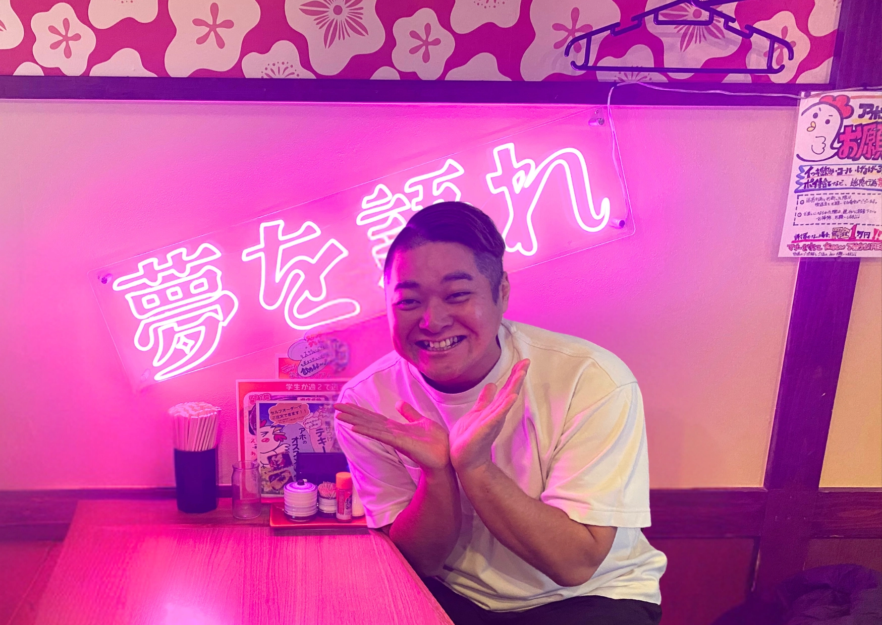 ピンクのネオンサイン「夢を語れ」の前で、白いTシャツ姿の男性が両手を頬の横に添えて笑っている